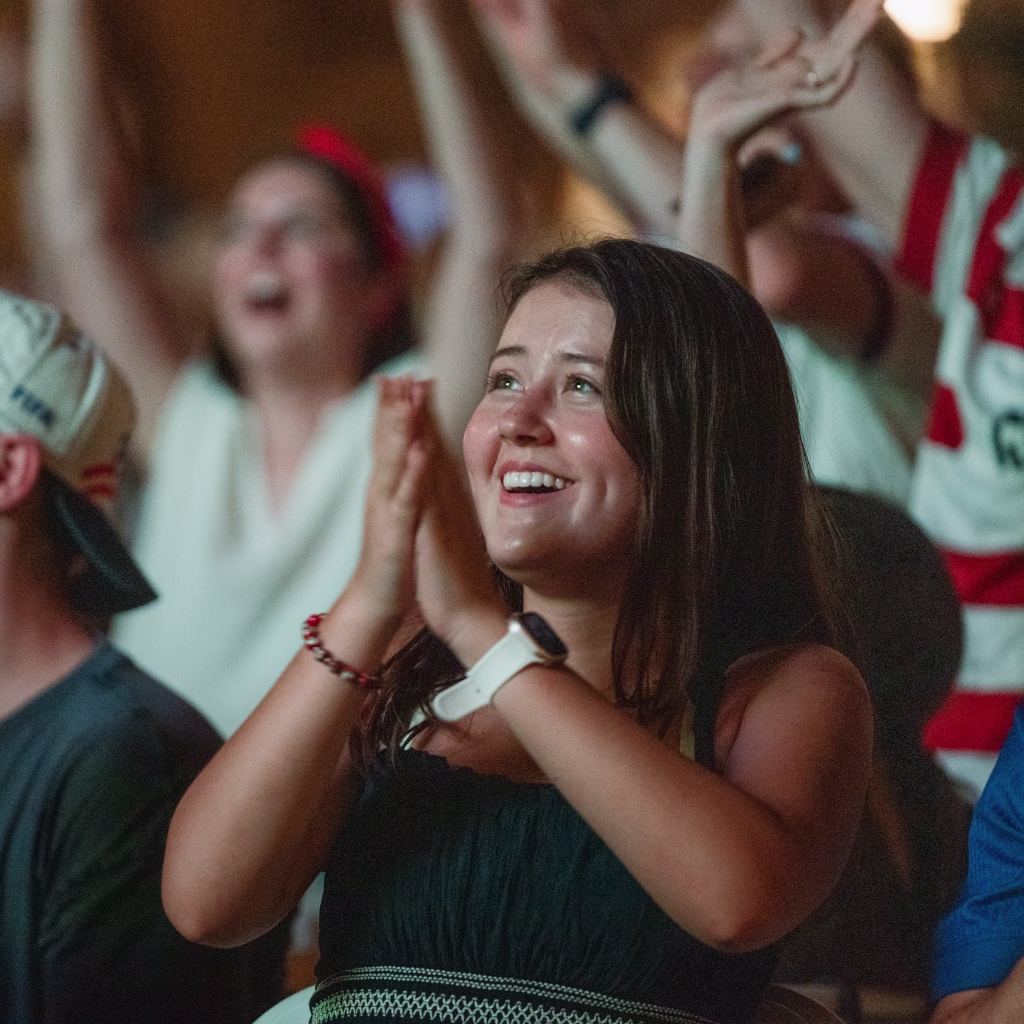 : Young woman smiling and clapping among a cheering crowd, suggesting excitement and celebration at a sports event.