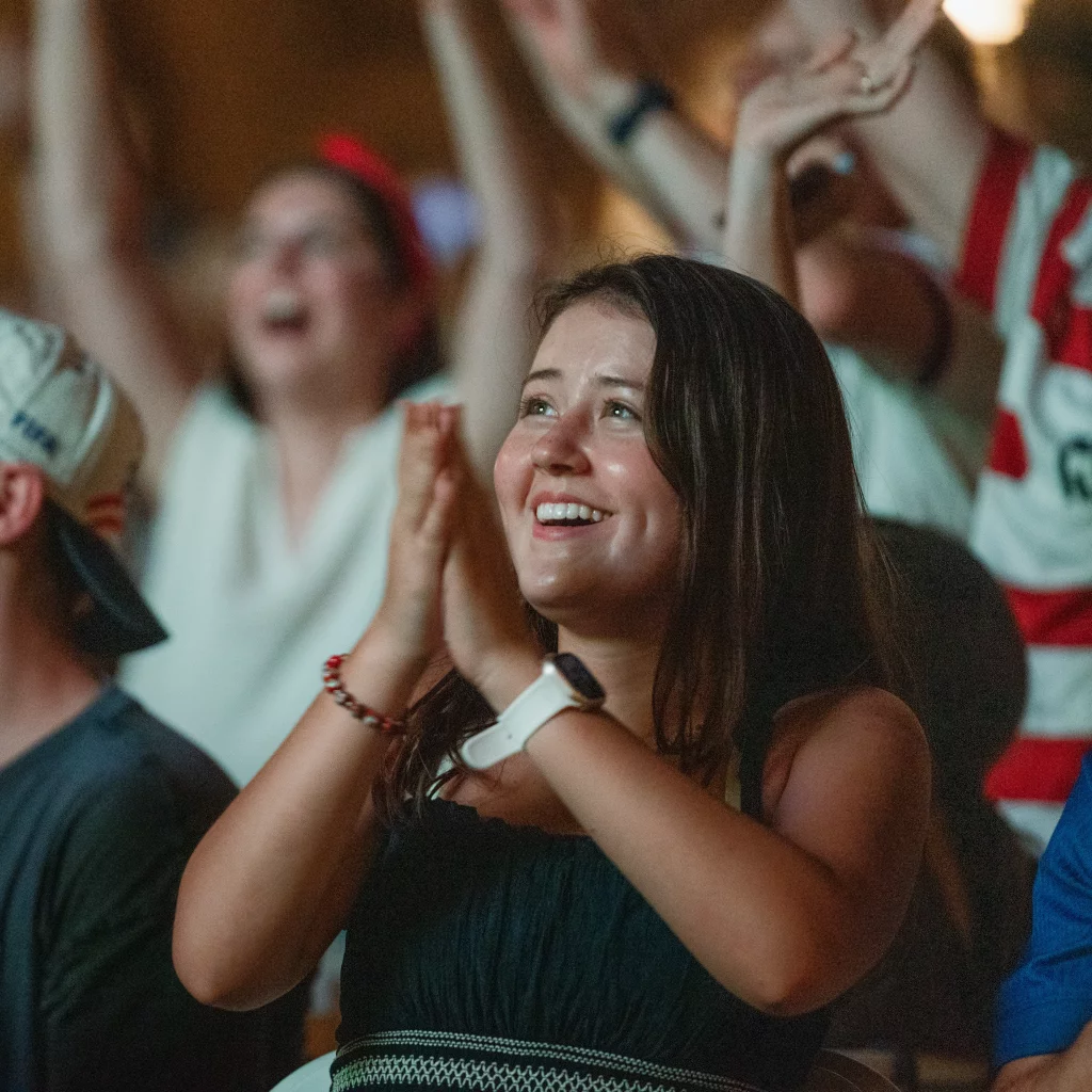 : Young woman smiling and clapping among a cheering crowd, suggesting excitement and celebration at a sports event.