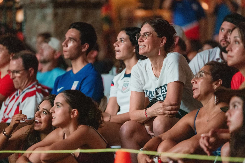 : A diverse group of people sit closely together, watching an event intently. Many wear United States soccer jerseys and shirts, suggesting they are fans at a World Cup viewing. Their expressions range from excited to anxious.