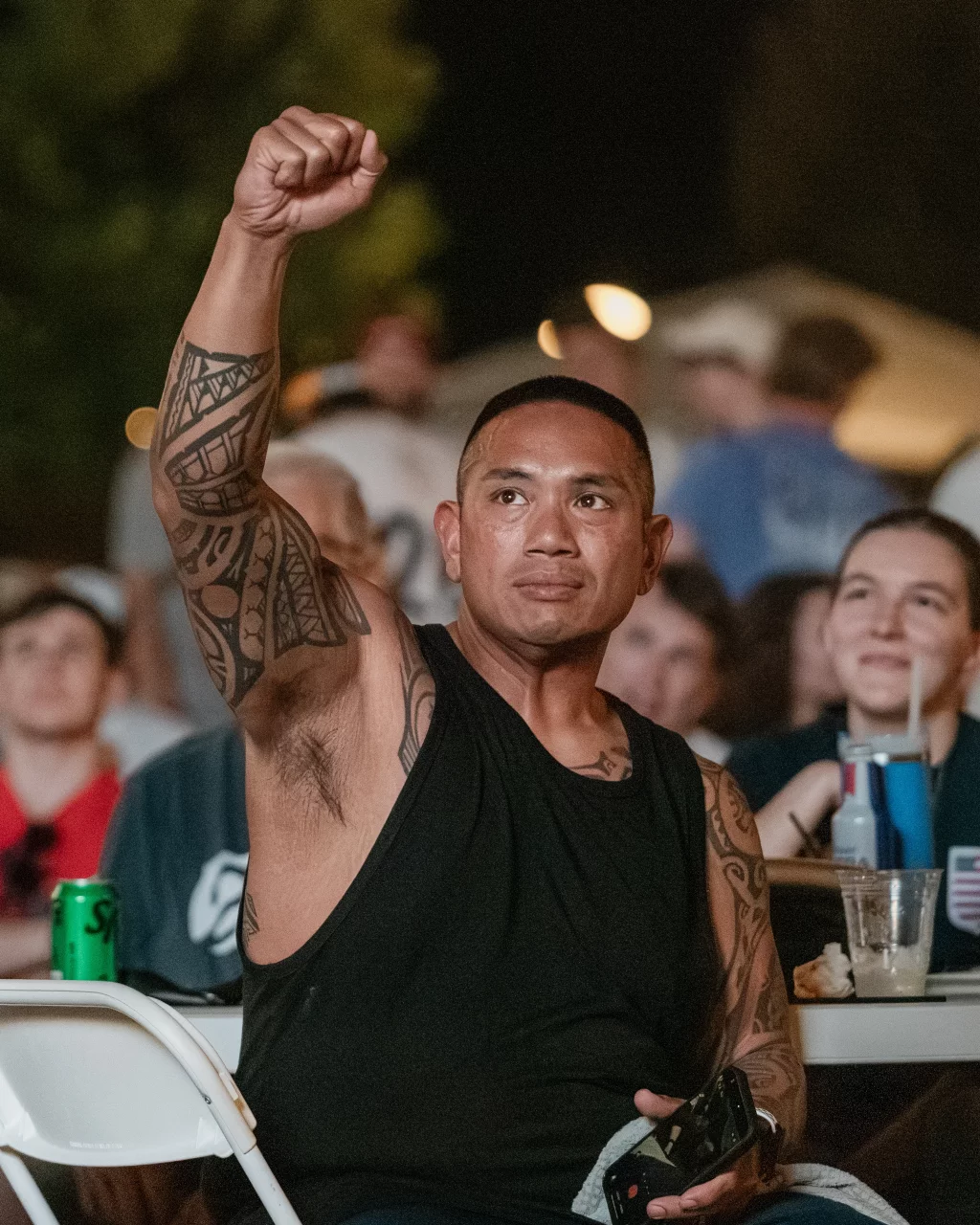 : Man with tattooed arm raises fist in the air while sitting among a crowd, appearing to celebrate or show support at an event.