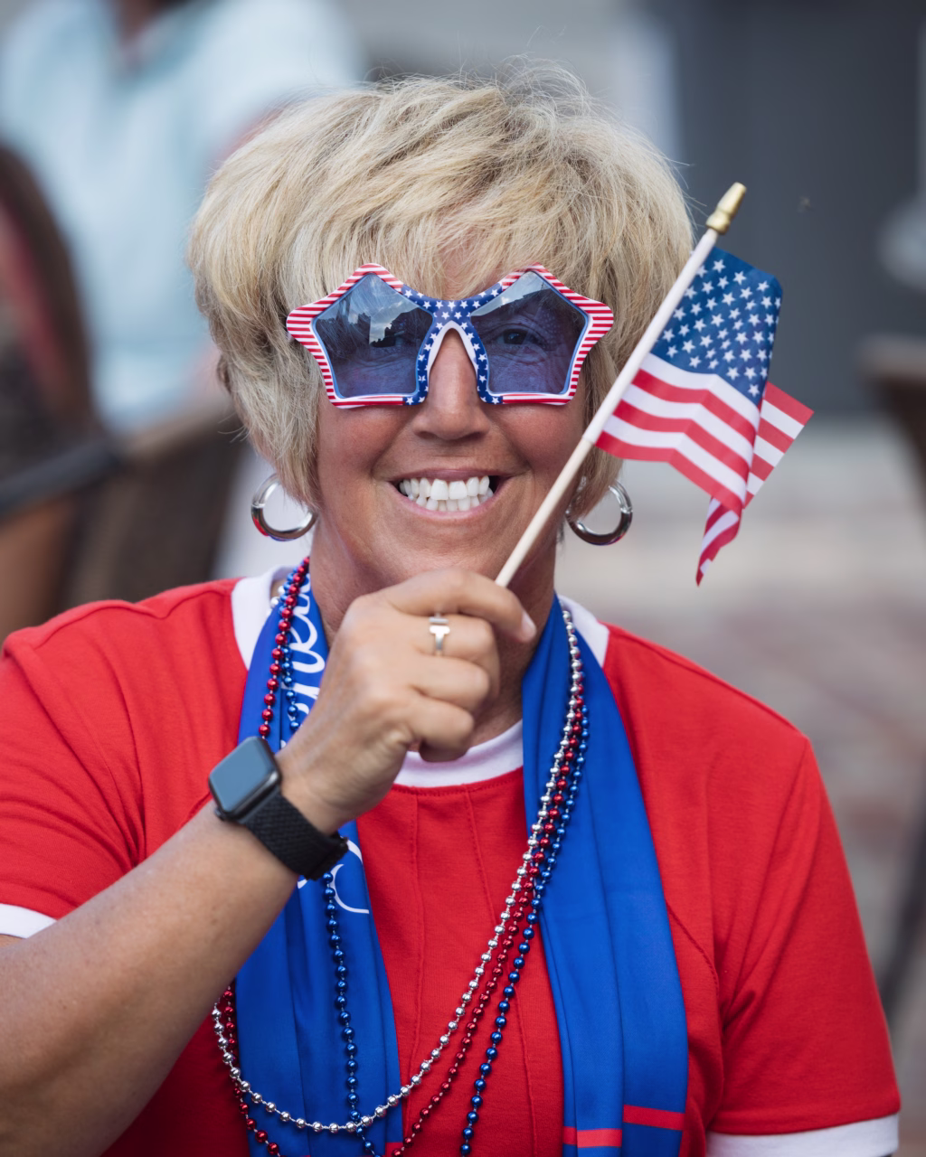 : Smiling person wearing star-shaped American flag sunglasses and red, white, and blue accessories, holding a small U.S. flag.
