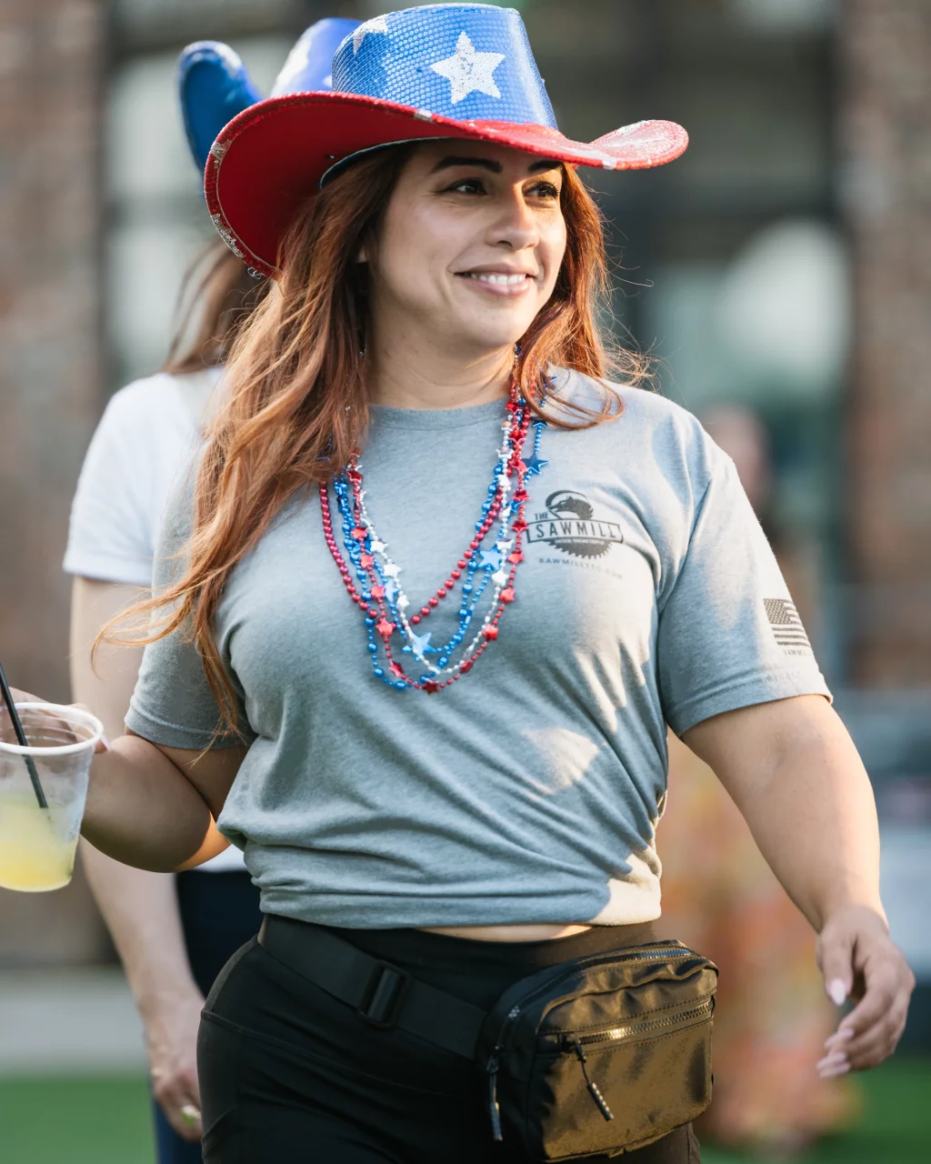 : Woman wearing a sparkly red, white, and blue cowboy hat, red and blue bead necklaces, a gray T-shirt, and a black fanny pack, holding a drink and smiling outdoors.