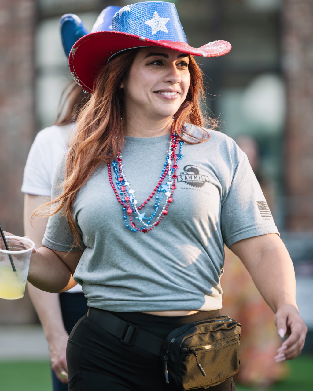: Woman wearing a sparkly red, white, and blue cowboy hat, red and blue bead necklaces, a gray T-shirt, and a black fanny pack, holding a drink and smiling outdoors.