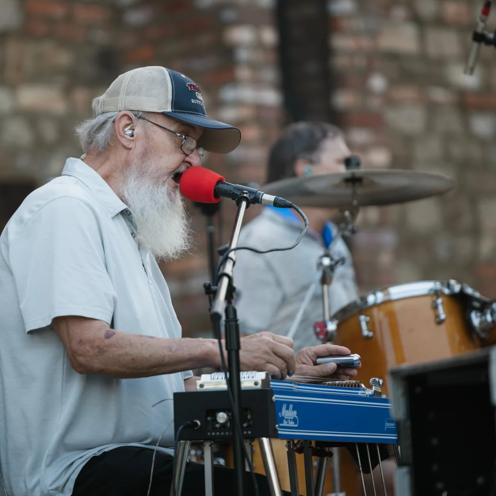 : An older man with a white beard and glasses plays a blue pedal steel guitar and sings into a red microphone, with a drum set and another musician in the background.
