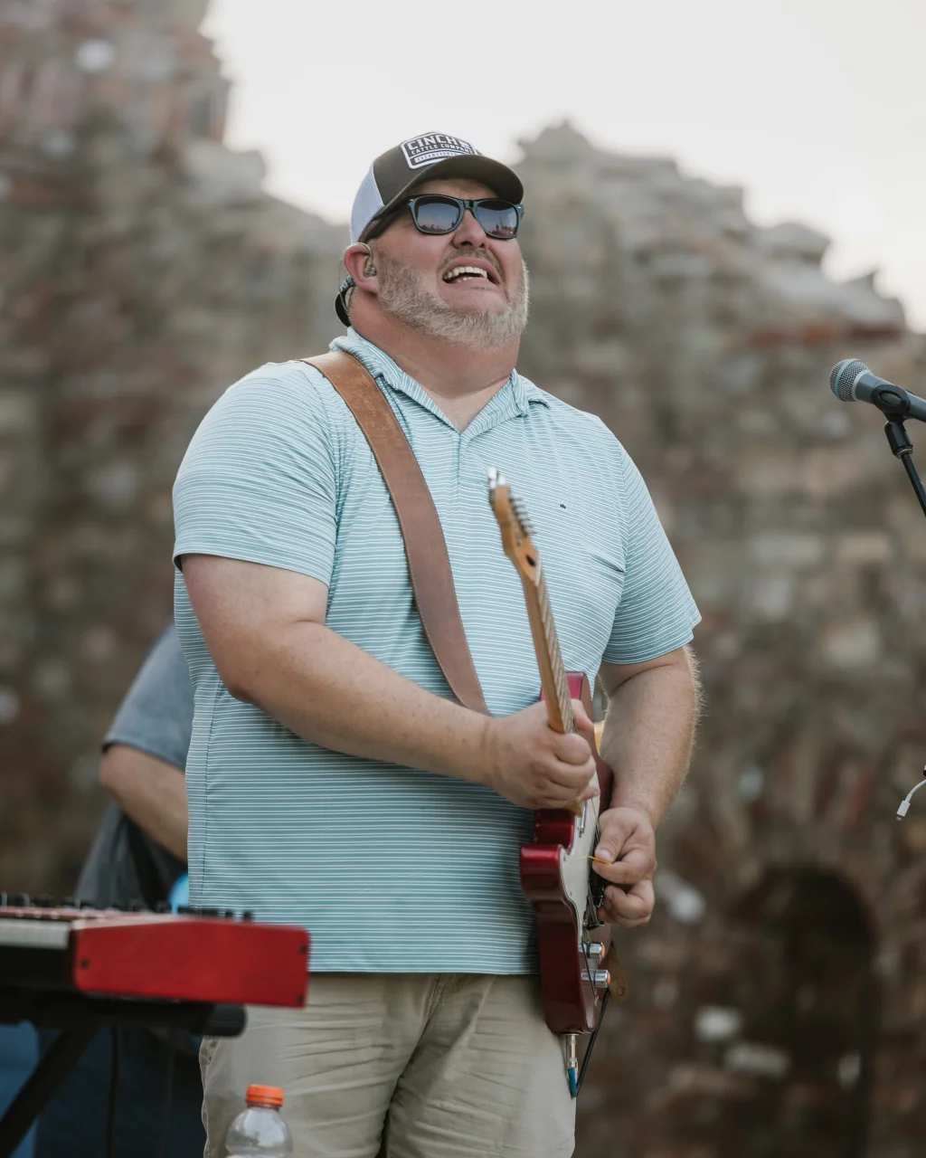 : Man wearing sunglasses and a baseball cap playing an electric guitar on an outdoor stage, with a microphone and keyboard visible nearby.