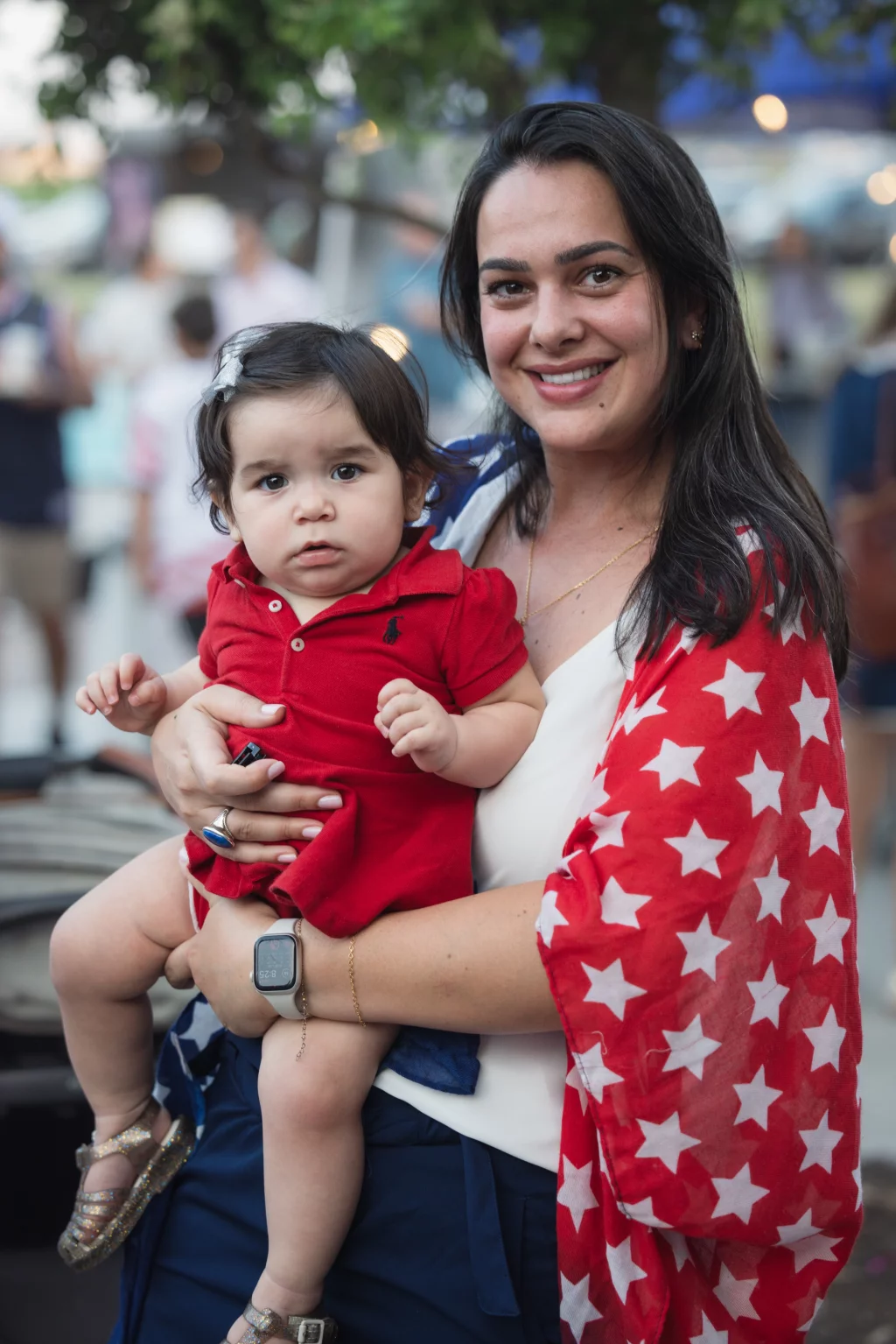 : Woman smiling and holding a young child dressed in a red outfit. The woman wears a red scarf with white stars and stands outdoors with a blurred crowd in the background.