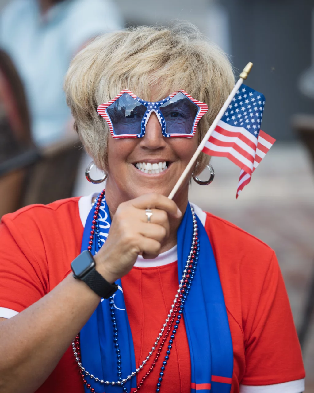 : Smiling person wearing red, white, and blue star-shaped sunglasses and holding a small American flag, dressed in festive attire with red, white, and blue beads and a blue scarf.