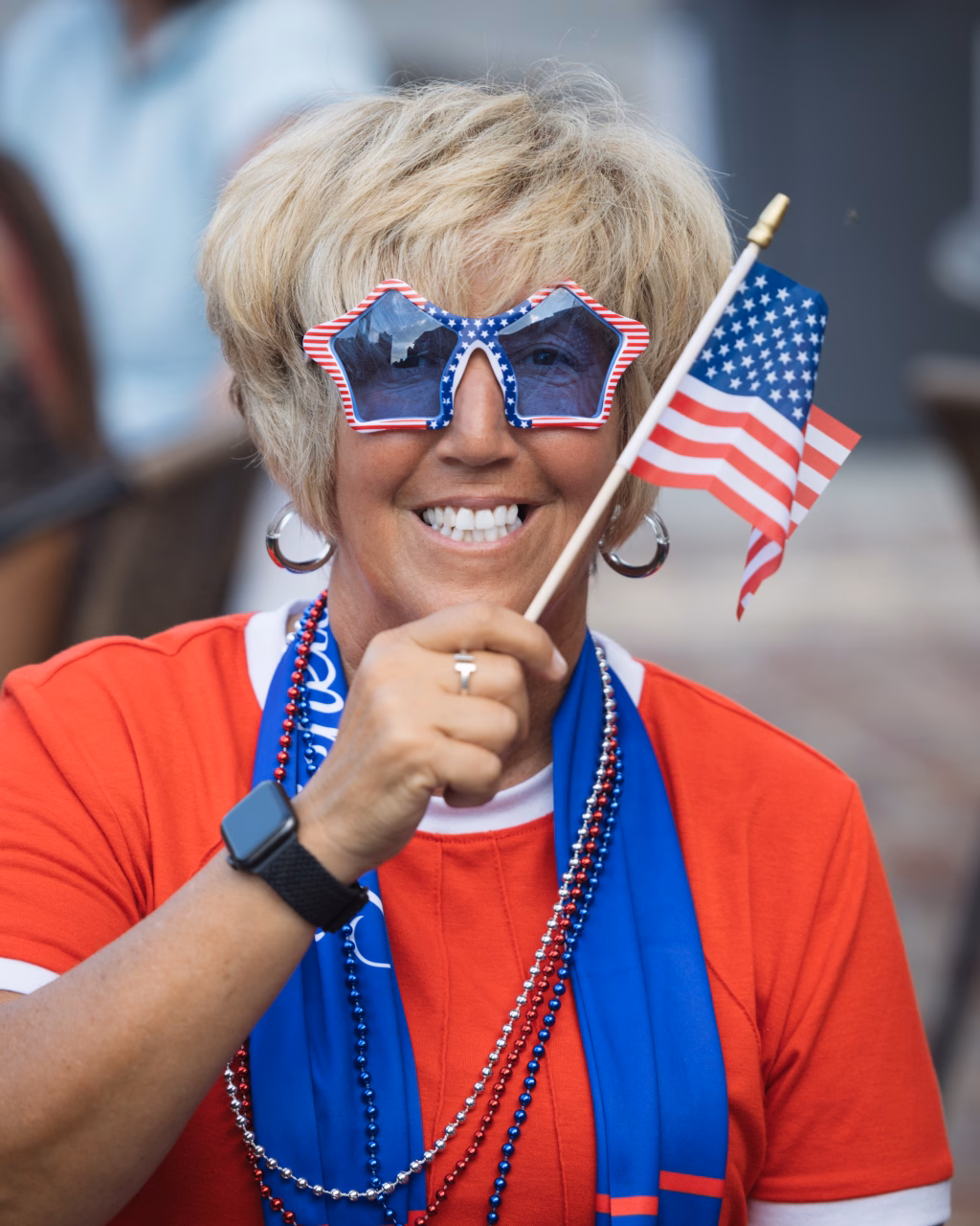 : Smiling person wearing red, white, and blue star-shaped sunglasses and holding a small American flag, dressed in festive attire with red, white, and blue beads and a blue scarf.