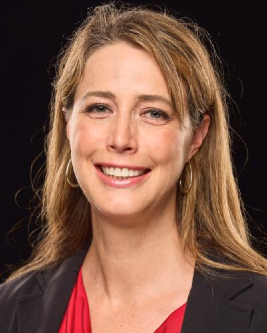 : Headshot of a smiling woman with long light brown hair, wearing a black blazer and red top, set against a black background.