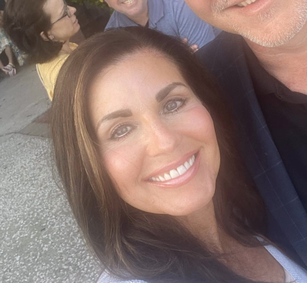 : Close-up selfie of a smiling woman with long dark hair and well-defined eyebrows, with a man partially visible next to her, outdoors on a paved surface with other people in the background.