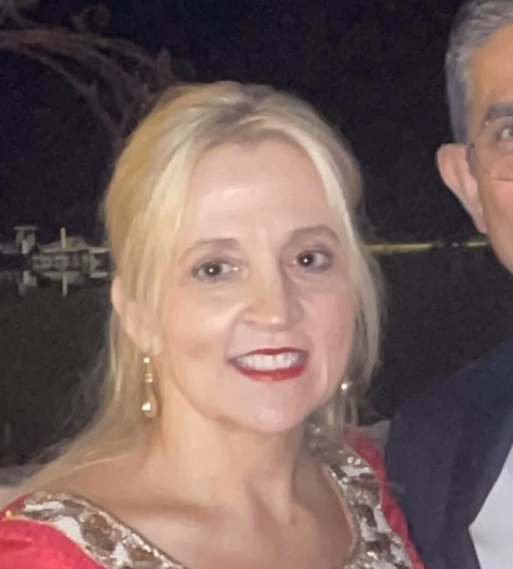 : Close-up of a smiling woman with blonde hair, wearing red lipstick, gold earrings, and a red and gold outfit, standing next to a man partially visible on the right, with a dark background.