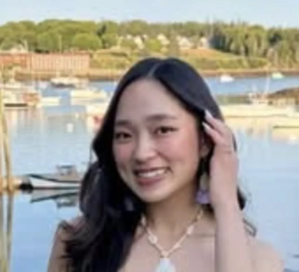 : A smiling woman with long dark hair poses by a sunlit lake, touching her hair, with trees and waterfront homes in the background.