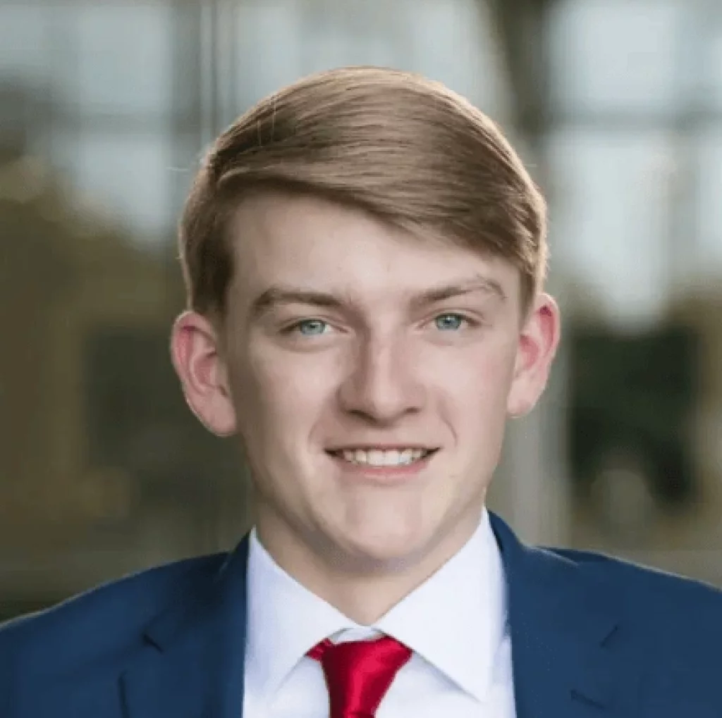 Alivia's senior photos taken Downtown, Greenville, SC: Young man with short light brown hair wearing a blue suit, white shirt, and red tie, smiling at the camera with a blurred indoor background.