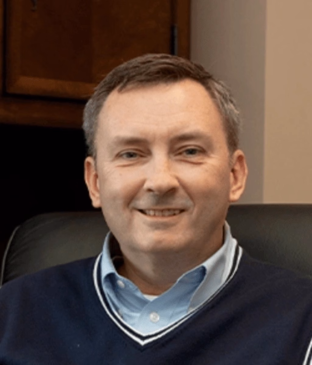 : A middle-aged man with short brown hair, wearing a navy blue sweater over a light blue collared shirt, sitting and smiling in an office setting.