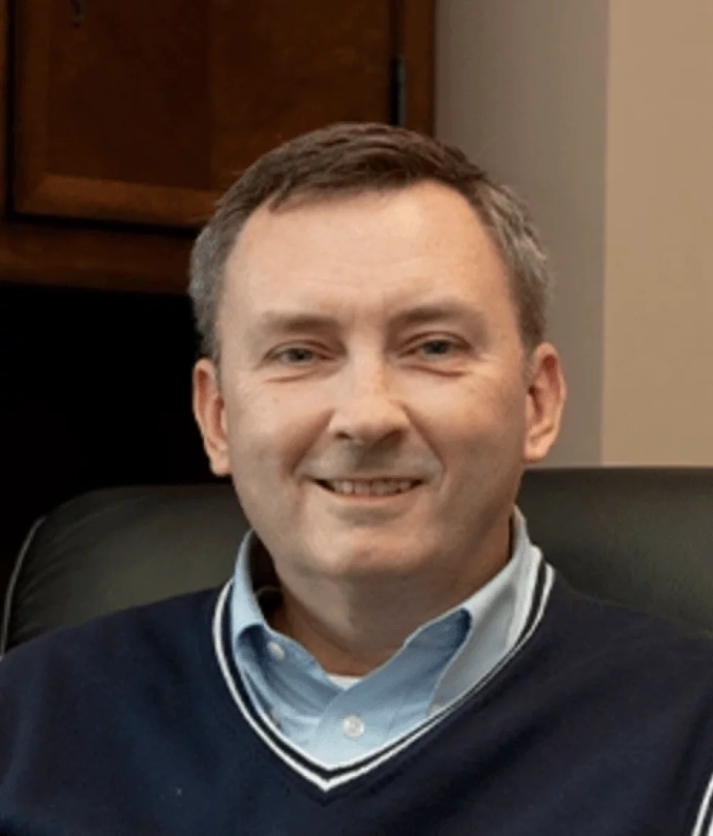 Alivia's senior photos taken Downtown, Greenville, SC: A middle-aged man with short brown hair, wearing a navy blue sweater over a light blue collared shirt, sitting and smiling in an office setting.