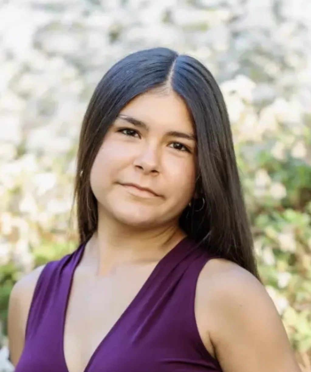 Olivia's senior photos taken at Furman University: A young woman with long straight dark hair, wearing a sleeveless purple top, stands outdoors in natural light with greenery and a blurred background behind her.