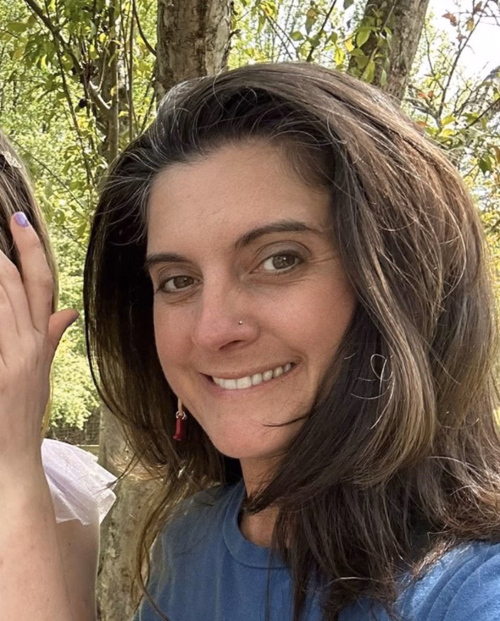 : Close-up of a smiling woman with long brown hair, wearing a blue shirt and a small nose stud, outdoors with trees in the background.